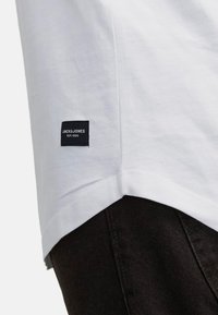T-shirt en coton blanc avec une étiquette noire portant l'inscription "JACK & JONES EST. 1990" sur la manche gauche, associé à un jean foncé.