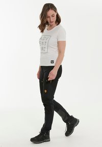 Vit t-shirt med grafisk tryck av "ADVENTURE" i svart, tillsammans med åtsittande svarta byxor med fickor och en beige logopatch. Svarta vandringskängor.