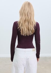 Langärmliges, figurbetontes Oberteil in dunklem Burgunderrot, aus weichem Stoff gefertigt; kombiniert mit hochgeschnittenen weißen Hosen mit Gesäßtaschen.