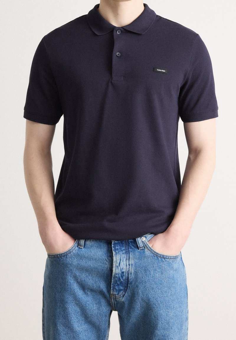 Personne portant un polo bleu marine à manches courtes avec un petit logo Calvin Klein et un jean bleu clair, mains dans les poches.