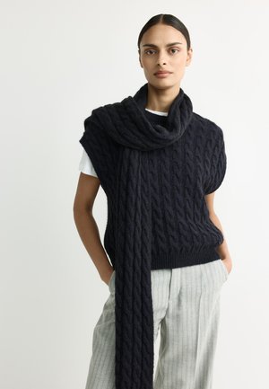Young woman with dark hair wears a black cable-knit sleeveless sweater and matching scarf, paired with light gray pinstriped trousers.
