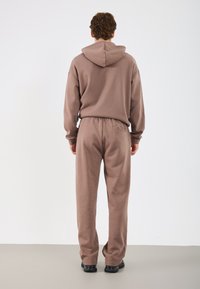 Brązowa bluza z kapturem i pasujące spodnie wykonane z miękkiego materiału, charakteryzujące się luźnym krojem i elastycznym pasem z detalem w postaci tylnej kieszeni.