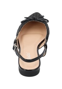 Chaussure noire tissée avec un accent de nœud et une sangle de cheville ajustable. Intérieur beige avec texture lisse et un talon solide bas.