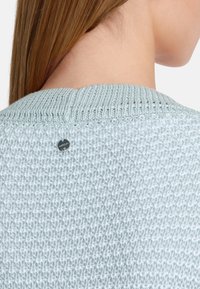 Hellblauer Strickpullover mit strukturiertem Muster. Er hat einen gerippten Kragen und ein kleines metallisches Logo-Detail auf der Rückseite.