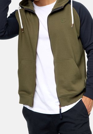 Mann trägt eine olivgrüne und schwarze Kapuzenjacke mit weißem Kordelzug über einem weißen T-Shirt und schwarzen Hosen, beschnitten an Hals und Oberschenkeln.