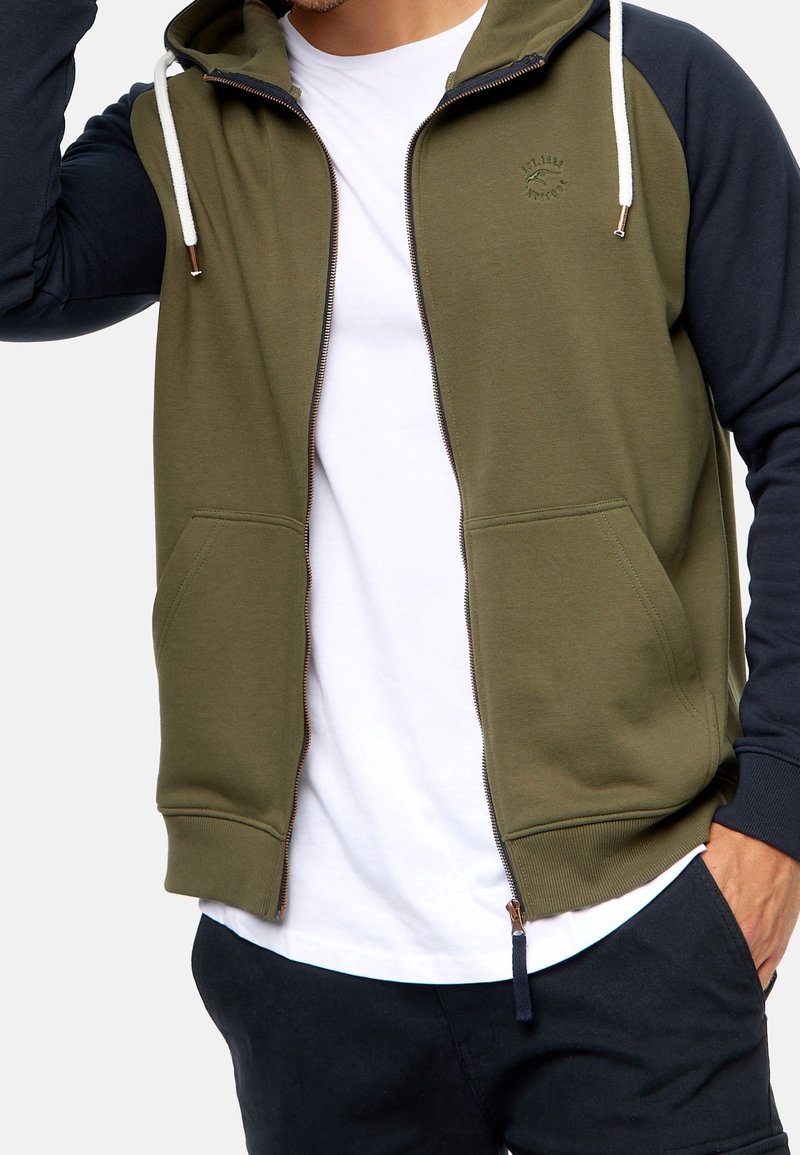 Homme portant un sweat à capuche zippé vert olive et noir avec des cordons blancs sur un t-shirt blanc et un pantalon noir, raccourci au niveau du cou et des cuisses.