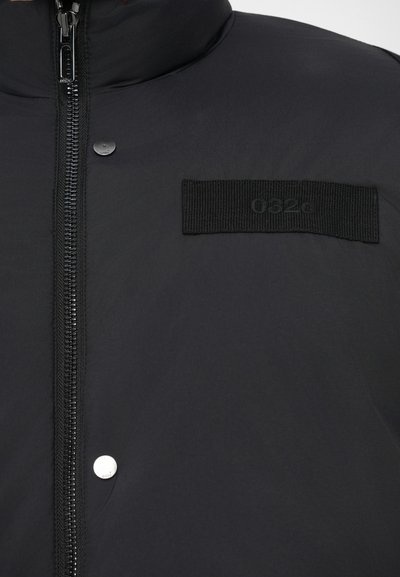 Czarna kurtka puchowa z zapięciem na zamek, detalami na napy oraz wyraźną czarną naszywką z logo "052g". Gładka tekstura.