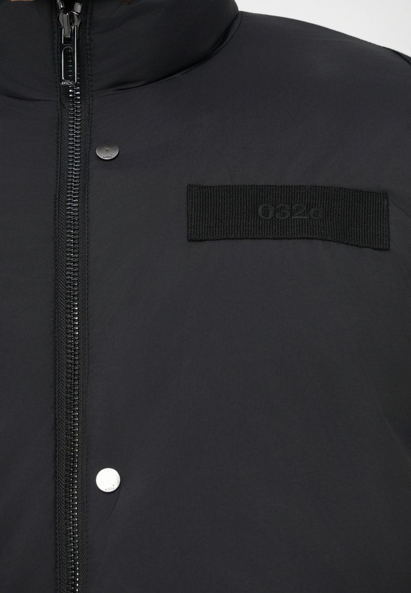 Schwarze Daunenjacke mit Reißverschluss, Druckknopfdetails und einem auffälligen schwarzen Stoffaufnäher mit dem "052g"-Logo. Glatte Textur.
