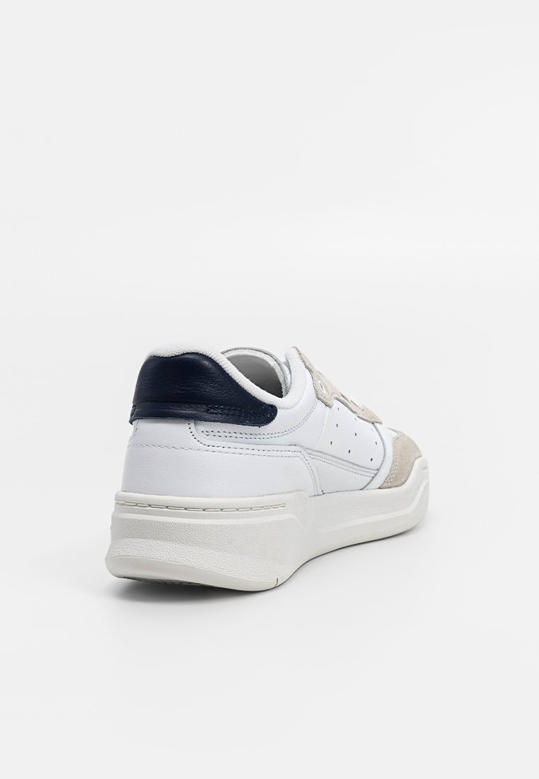 Zapatilla blanca con acentos en beige y marino, que presenta un upper de cuero liso, detalles de gamuza, un talón texturizado y un diseño de suela gruesa.