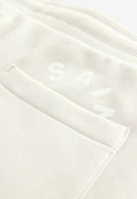 Světle béžová látková kapsa s matným povrchem; obsahuje text "SAIZ" v bílé barvě, umístěný diagonálně na kapse. Viditelné detaily texturovaného povrchu.