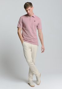 Lichtroze polo shirt met een kraag en drie knopen, gecombineerd met crèmekleurige broeken en lichtgrijze sneakers. Eenvoudig ontwerp, zonder patronen.