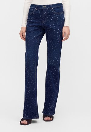 Jean en denim bleu foncé à coupe évasée, décoré de petites pois blancs, porté avec des sandales noires ouvertes et un haut blanc à manches longues.