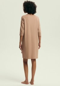 Kobieta z kręconymi włosami stoi boso, ubrana w luźną, beżową sukienkę do kolan z rękawami do trzech kwart, zwrócona plecami na jasnym tle.