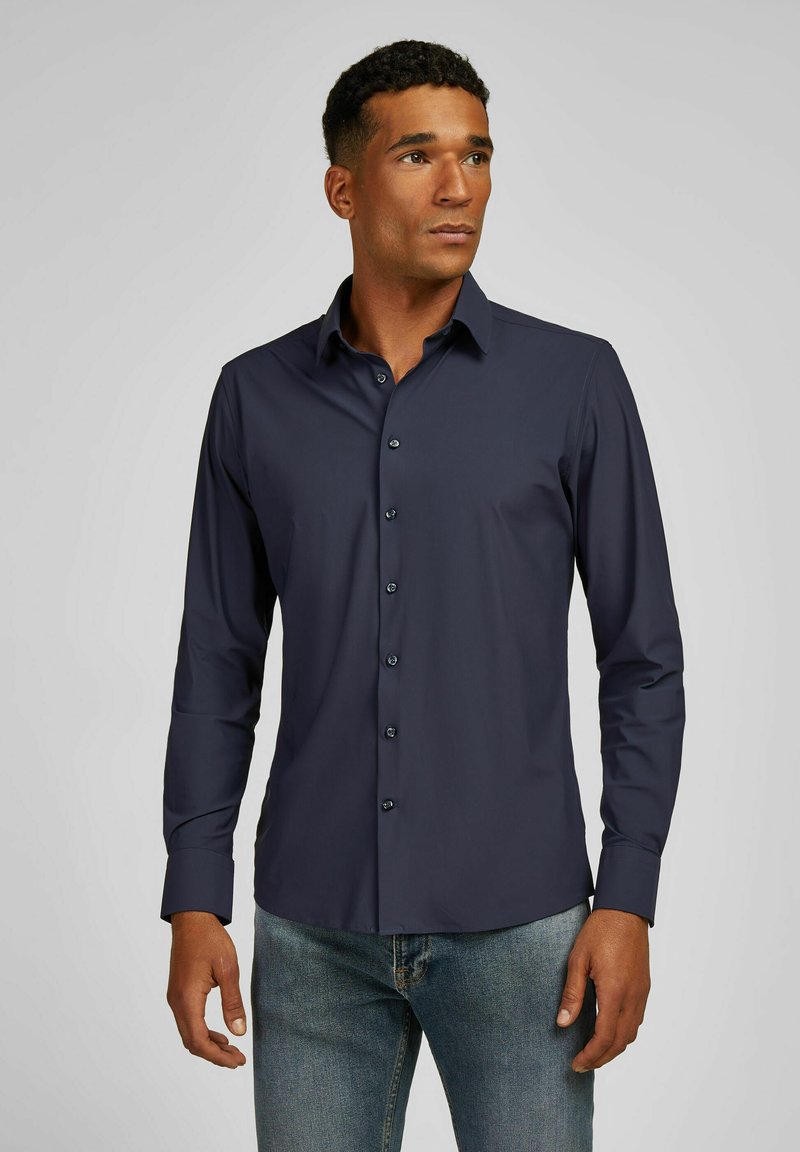 Marineblauw overhemd met lange mouwen en knoopsluiting, gemaakt van gladde stof, met gestructureerde kraag en bijpassende knopen. Gepaird met lichtblauwe jeans, die een getailleerd ontwerp tonen.