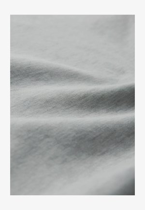 Närbild av mjuk, ljusgrå tyg med synlig fin vävstruktur och mjuka veck som skapar subtila skuggor och högdagrar.