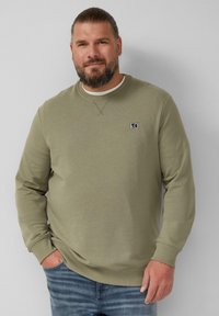 Felpa verde oliva con collo rotondo, polsini a coste e un piccolo logo sul petto; abbinata a jeans in denim di un lavaggio medio.