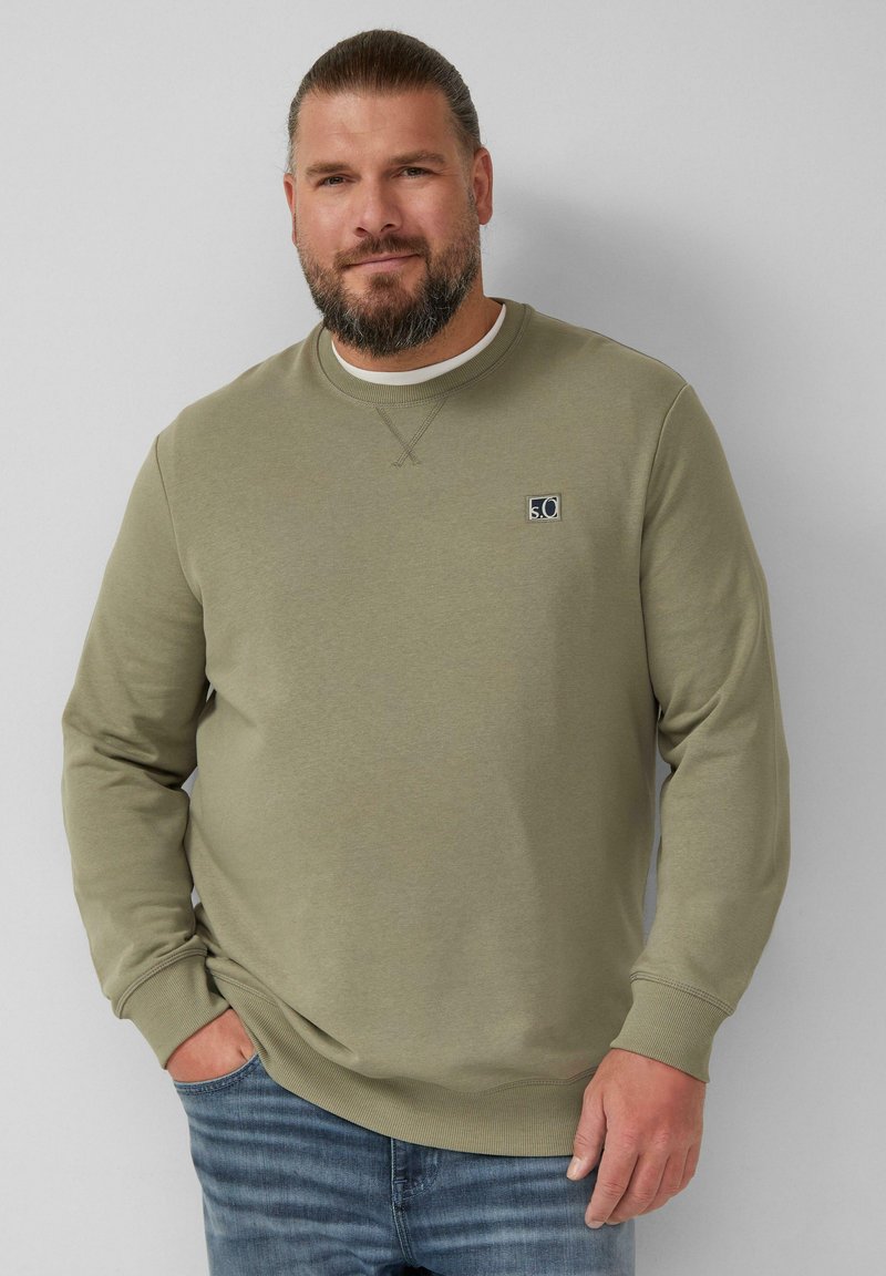 Felpa verde oliva con collo rotondo, polsini a coste e un piccolo logo sul petto; abbinata a jeans in denim di un lavaggio medio.