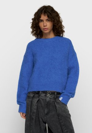Kvinde med krøllet hår iført en blå fluffy sweater og løse sorte vaskede jeans med bælte, stående mod en ensfarvet lys baggrund.