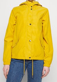 Żółta lekka kurtka z kapturem, zatrzaskami, dwoma bocznymi kieszeniami, elastycznymi mankietami i sznurkiem w dolnym obszyciu. Gładka faktura i casualowy design.