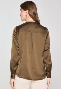 Camicia lunga in satin marrone con yoke posteriore pulito, dettaglio di pieghe sottili e polsini con bottoni. Texture liscia e vestibilità su misura.