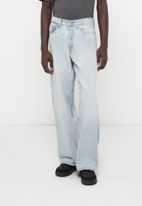 Jeans de pierna ancha de color azul claro con un acabado desgastado, cinco bolsillos y cierre de botones. Combinados con zapatos negros y una camiseta gris.