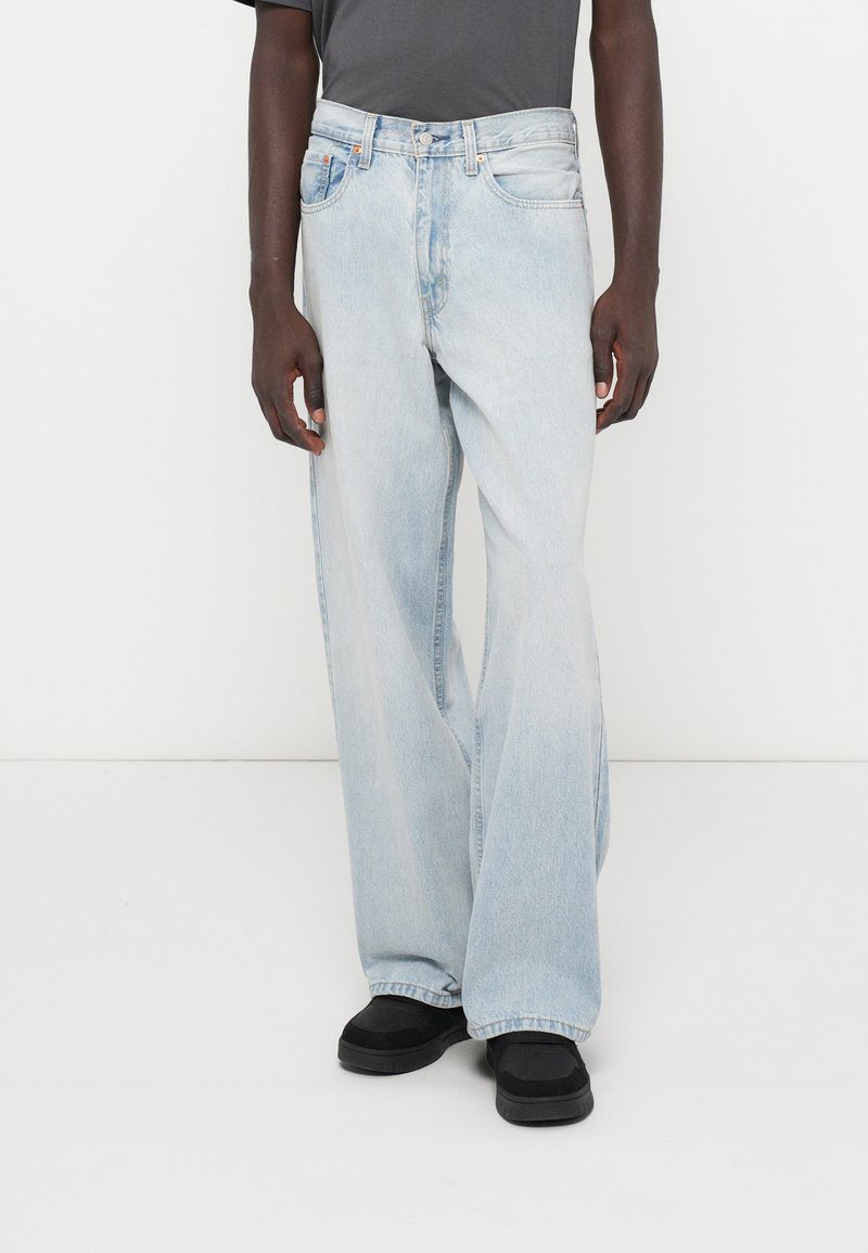 Jeans de pierna ancha de color azul claro con un acabado desgastado, cinco bolsillos y cierre de botones. Combinados con zapatos negros y una camiseta gris.