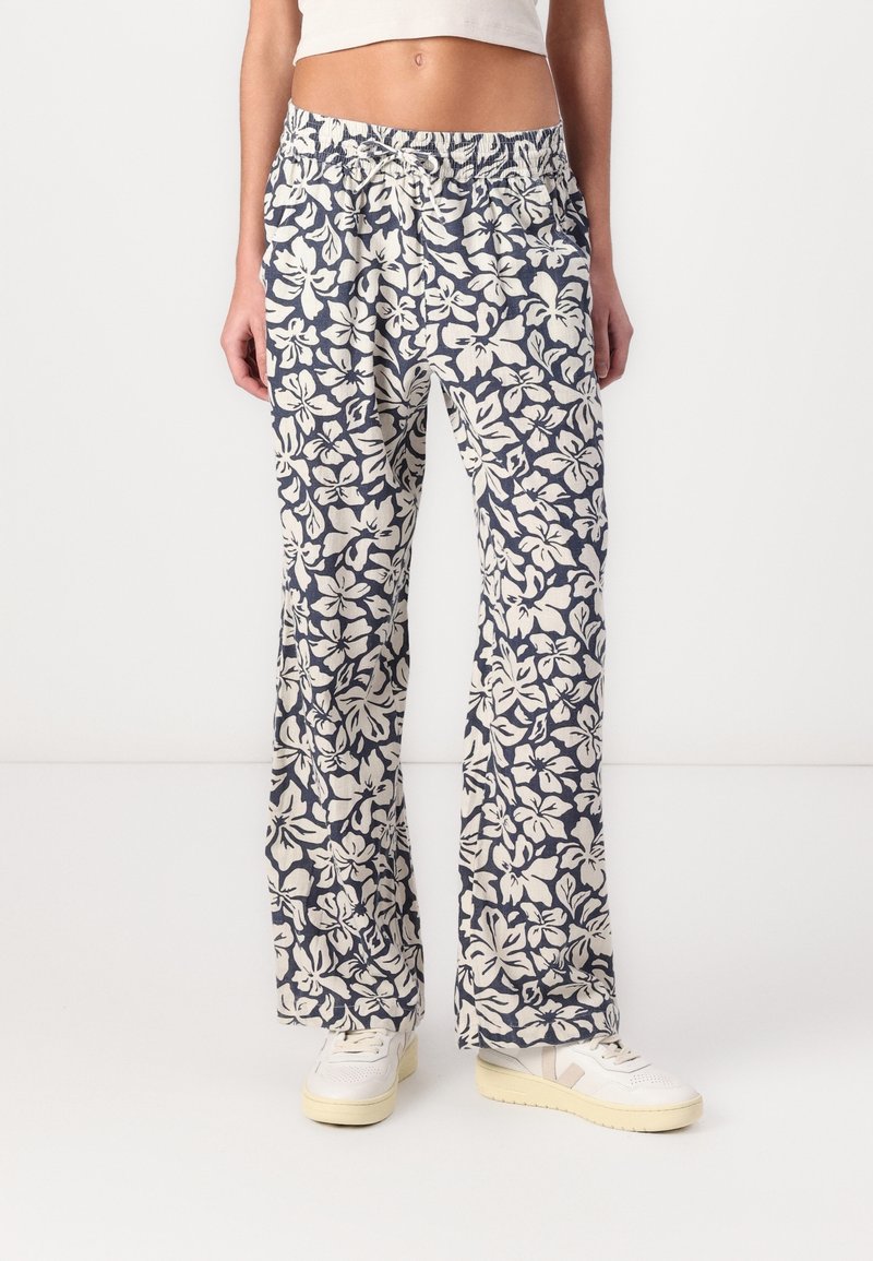 Personne portant un pantalon ample taille haute avec un motif floral blanc et bleu foncé, associé à des baskets blanches sur un fond uni.