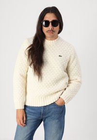 Cream-colored knitted sweater with a textured pattern and ribbed cuffs, worn with blue jeans. Features a green Lacoste logo on the chest.