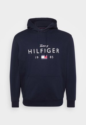 Dunkelblauer Kapuzenpullover aus weichem Stoff, mit einem großen weißen "Hilfiger"-Logo, einem kleineren "Tommy" darüber und einem Farbblockakzent in Rot und Weiß.