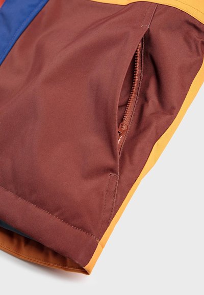 Un tissu brun avec une texture lisse présente une poche zippée. Le vêtement a des accents jaunes et bleus le long des bords.