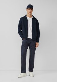 Dunkelblaue Reißverschlussjacke über einem weißen T-Shirt, kombiniert mit dunkelblauen Hosen und weißen Turnschuhen. Das Modell trägt eine passende dunkelblaue Kappe.