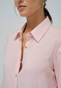 Camisa de color rosa claro con cuello y diseño de botones, que presenta detalles de herrajes dorados, fabricada en un tejido ligero y de textura suave.