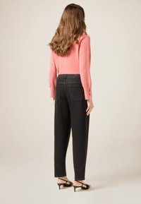 Mujer con cabello largo y castaño, vestida con un suéter rosa, jeans negros de talle alto y sandalias negras de tacón bajo, posando frente a un fondo liso.