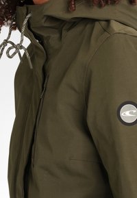 Veste imperméable verte avec fermeture éclair, capuche réglable, accents à cordon et un écusson circulaire sur la manche gauche.