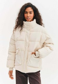 Béžová prošívaná péřová bunda s vysokým límcem, dvěma předními kapsami a manžetovými rukávy. Měkká textura s jemným vzorem detailů.