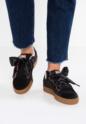 Osoba nosząca czarne buty Puma z grubymi sznurowadłami z czarnej satyny i gumową podeszwą, w połączeniu z niebieskimi dżinsami z postrzępionym wykończeniem.