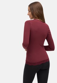 Camisa burdeos de manga larga con detalle de encaje en el borde. La tela parece suave y ajustada, combinada con leggings negros.