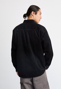 Un homme portant une chemise en velours côtelé noire et un pantalon cargo gris se tient de profil devant un fond blanc uni.