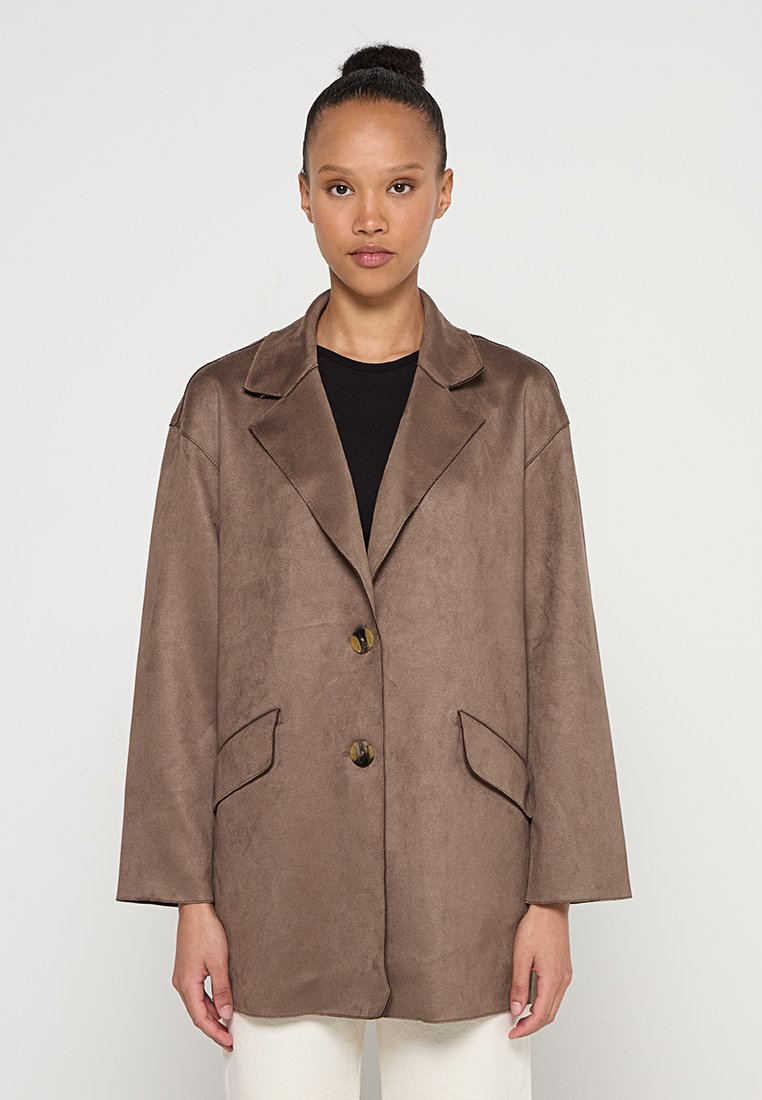 Donna con i capelli raccolti in un bun, che indossa un blazer oversize marrone su una maglietta nera e pantaloni bianchi, in piedi di fronte a uno sfondo chiaro e semplice.