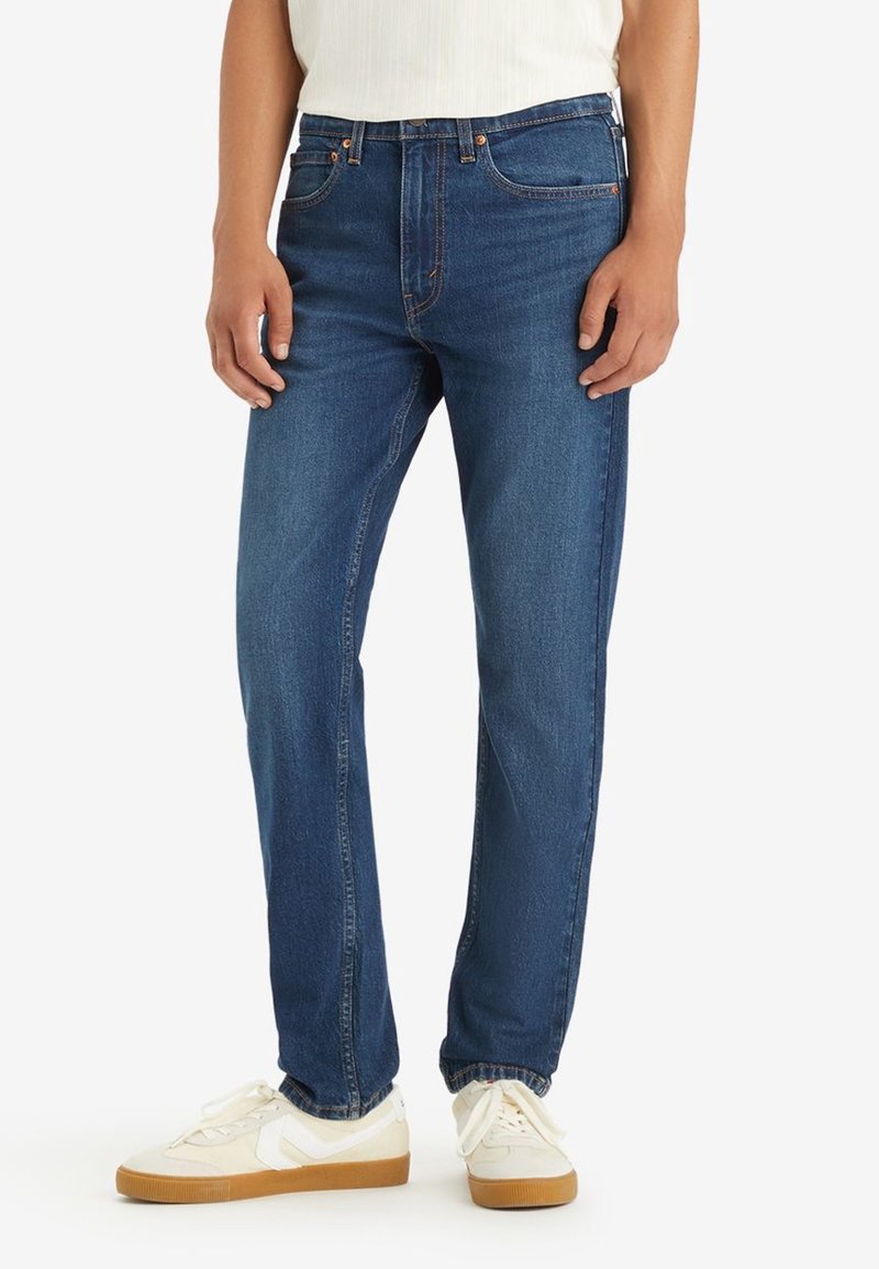 Jean en denim bleu foncé avec une coupe droite, comportant cinq poches et des détails de couture discrets. Portés avec des baskets blanches.