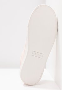 Světle růžová plátěná teniska s bílou gumovou podrážkou, s texturovaným dnem a vyraženým logem "Polo". Boční pohled, opřená o zeď.