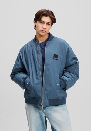 Blauwe bomberjack met een rits aan de voorkant, geribbelde manchetten en een klein zwart logo patch. Heeft een gladde textuur en een ontspannen pasvorm.