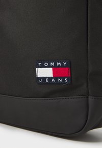 Svart syntetisk ryggsäck med en slät botten. Har en flerfärgad broderad logotyp "TOMMY JEANS" på en texturerad panel.