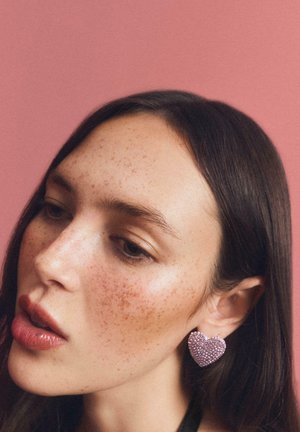 Szív alakú fülbevalók rózsaszín strasszkövekkel borítva. A dizájn merész, fényes textúrája kontrasztban áll a lágyan megvilágított háttérrel.