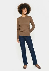 Brauner Strickpullover mit Rundhalsausschnitt und langen Ärmeln, kombiniert mit dunkelblauen Jeans und Schuhen im Tiermuster. Glatte Textur, taillierte Passform.