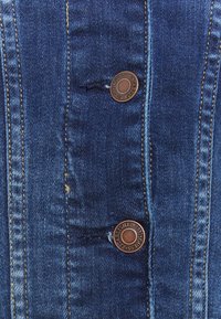 Blå denimjacka med kopparfärgad knappar, framträdande sömmar, en v distressed textur med små trasningar och fem fickor.