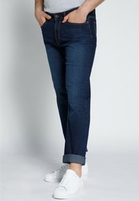 Jeans in denim blu scuro con una vestibilità slim, dotati di un classico design a cinque tasche, orli arrotolati e dettagli di cucitura a contrasto. Indossati con sneakers bianche.