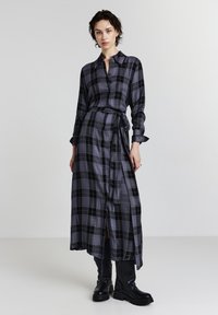 Donna che indossa un vestito camicia a quadri grigio e nero con bottoni, una cintura in vita e stivali neri fino al ginocchio, in piedi su uno sfondo bianco semplice.