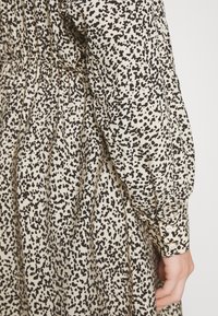 Vestido de tecido bege com padrão salpicado em preto, apresentando mangas longas, cintura elástica e detalhes em botão nos punhos.