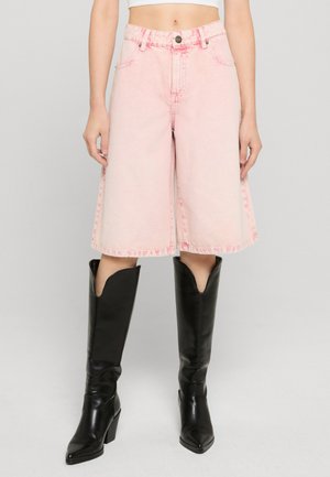 Kvinde iført højttaljede, lyserøde denimshorts til knæet kombineret med sorte knælange støvler med hæl på neutral baggrund.